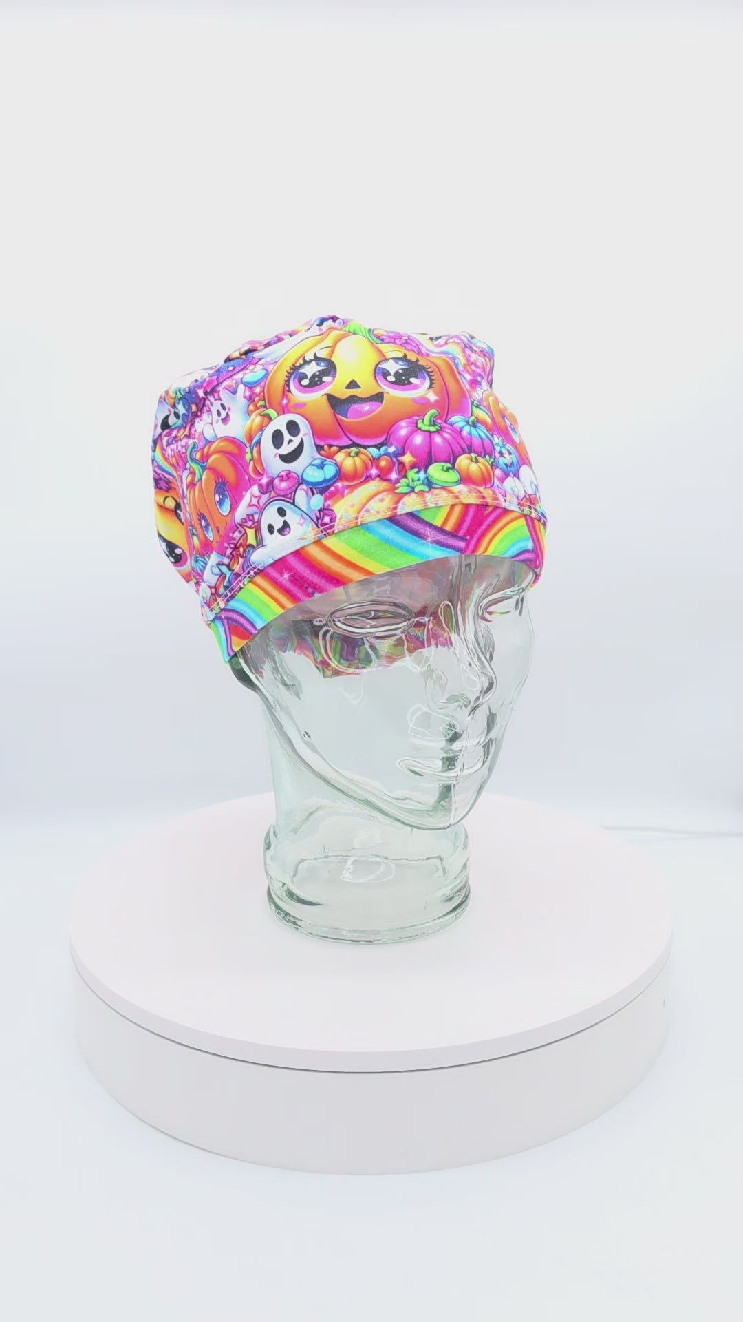 360-degree rotating video display of a full-coverage medical scrub cap featuring a colorful rainbow pumpkin pattern with cute jack-o'-lantern faces on a glass mannequin head.